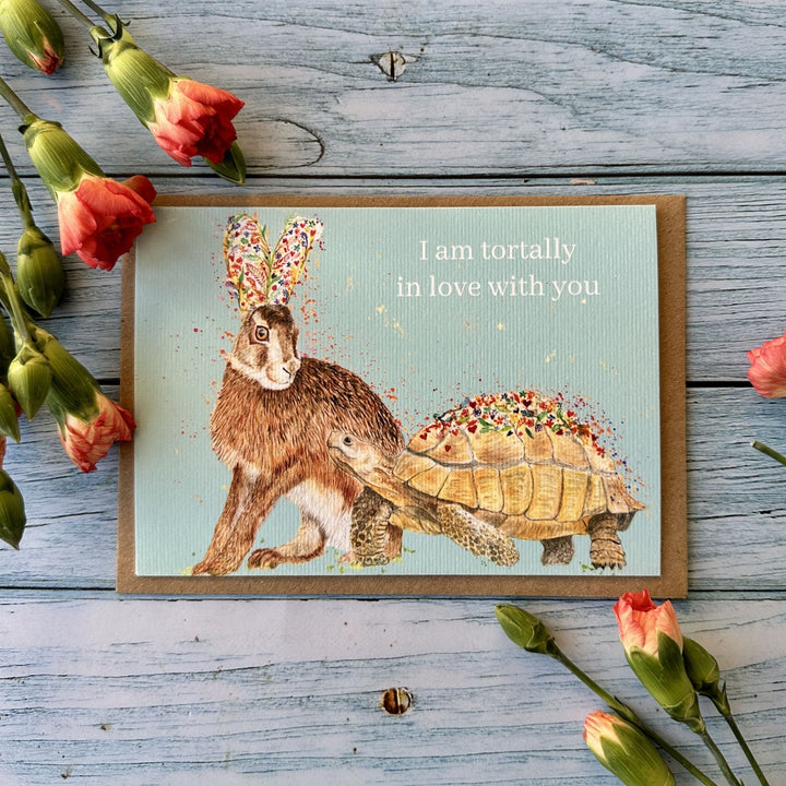 A square greeting card with a brown kraft border, featuring a watercolor-style hare and tortoise on a pale blue background with the text &