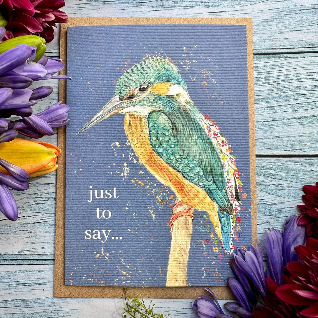 A square greeting card featuring a painted kingfisher on a blue-gray background with the text 'just to say.